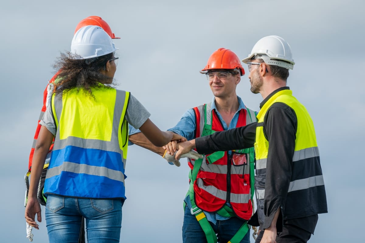 Four construction workers from Revival Construction, wearing safety vests and helmets, stand outdoors and join hands in the center as a team gesture under a cloudy sky.