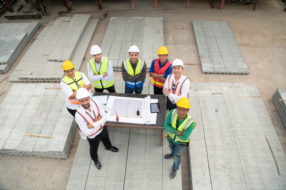 A group of six builders and engineers wearing safety vests and helmets stand around a table with blueprints at a construction site, looking up and smiling at the camera—ready to take on the next home renovation project.