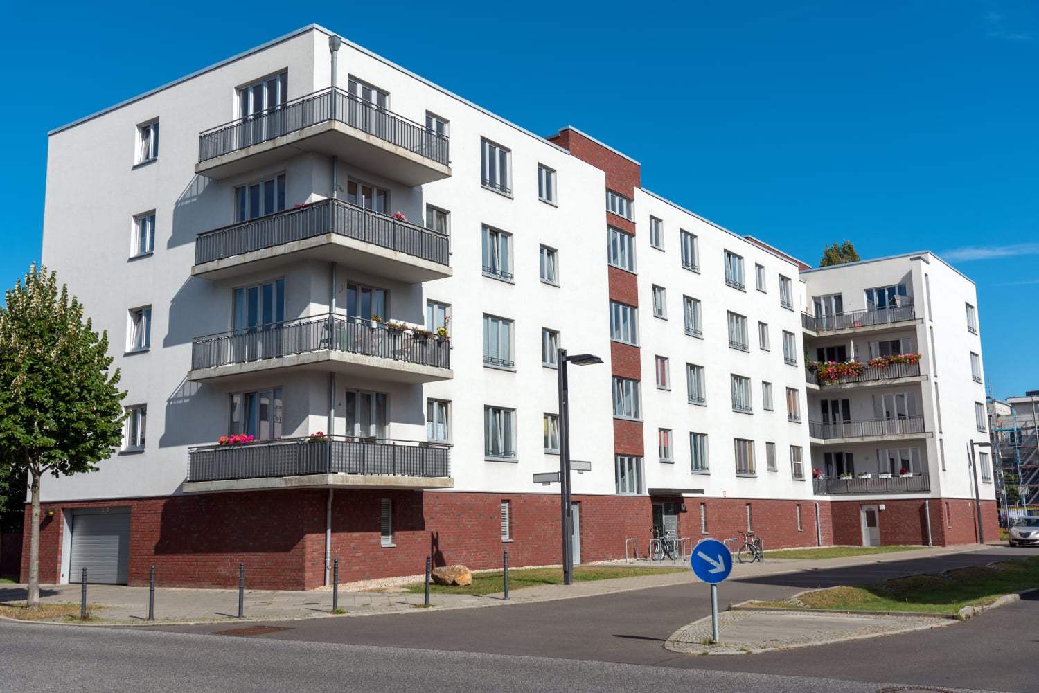 A modern four-story apartment building with white walls and red brick accents, expertly crafted by Revival Construction, featuring balconies and large windows, situated on a corner with a blue sky in the background.