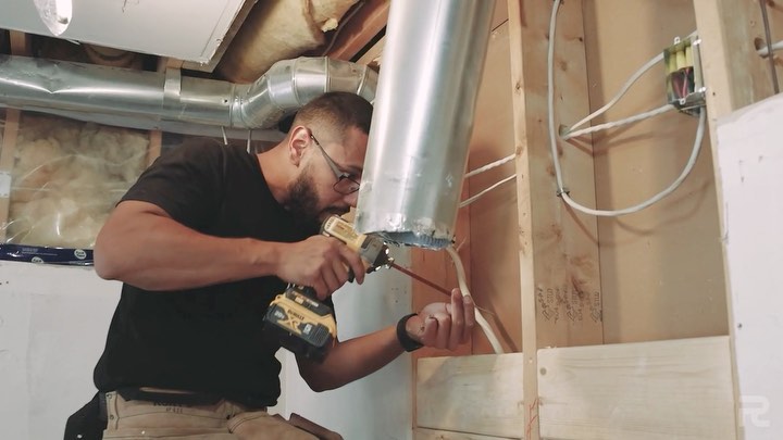 A man wearing glasses uses a power drill to install ductwork in a partially finished room with exposed studs and insulation, showcasing the expertise of a home renovation contractor from Revival Construction.