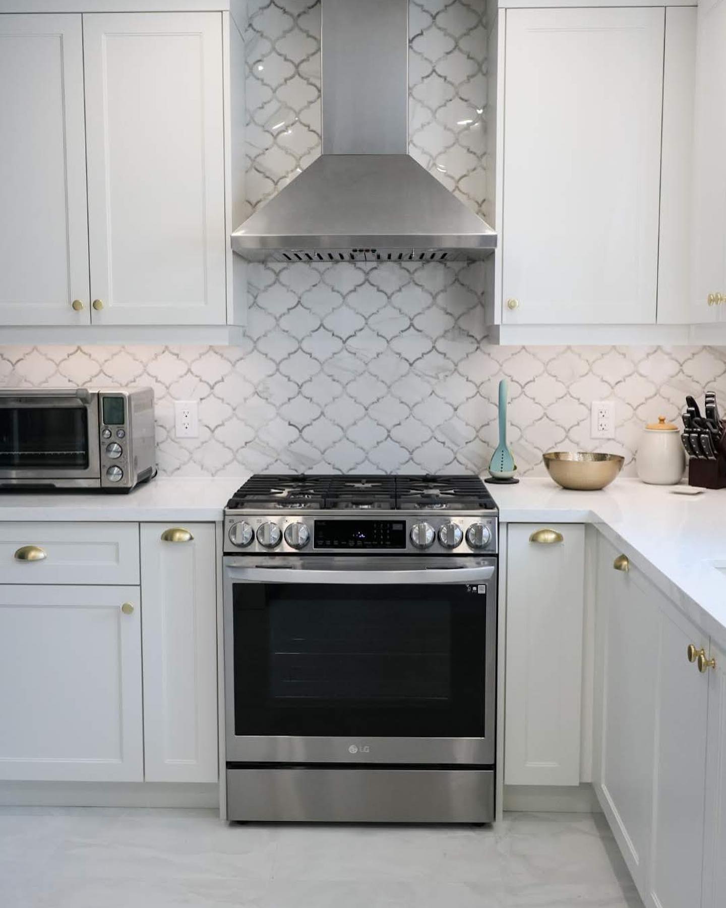 A modern kitchen by Revival Construction features white cabinets with gold handles, a stainless steel oven and range hood, a patterned tile backsplash, toaster oven, mixing bowl, utensils, and a sleek white countertop.