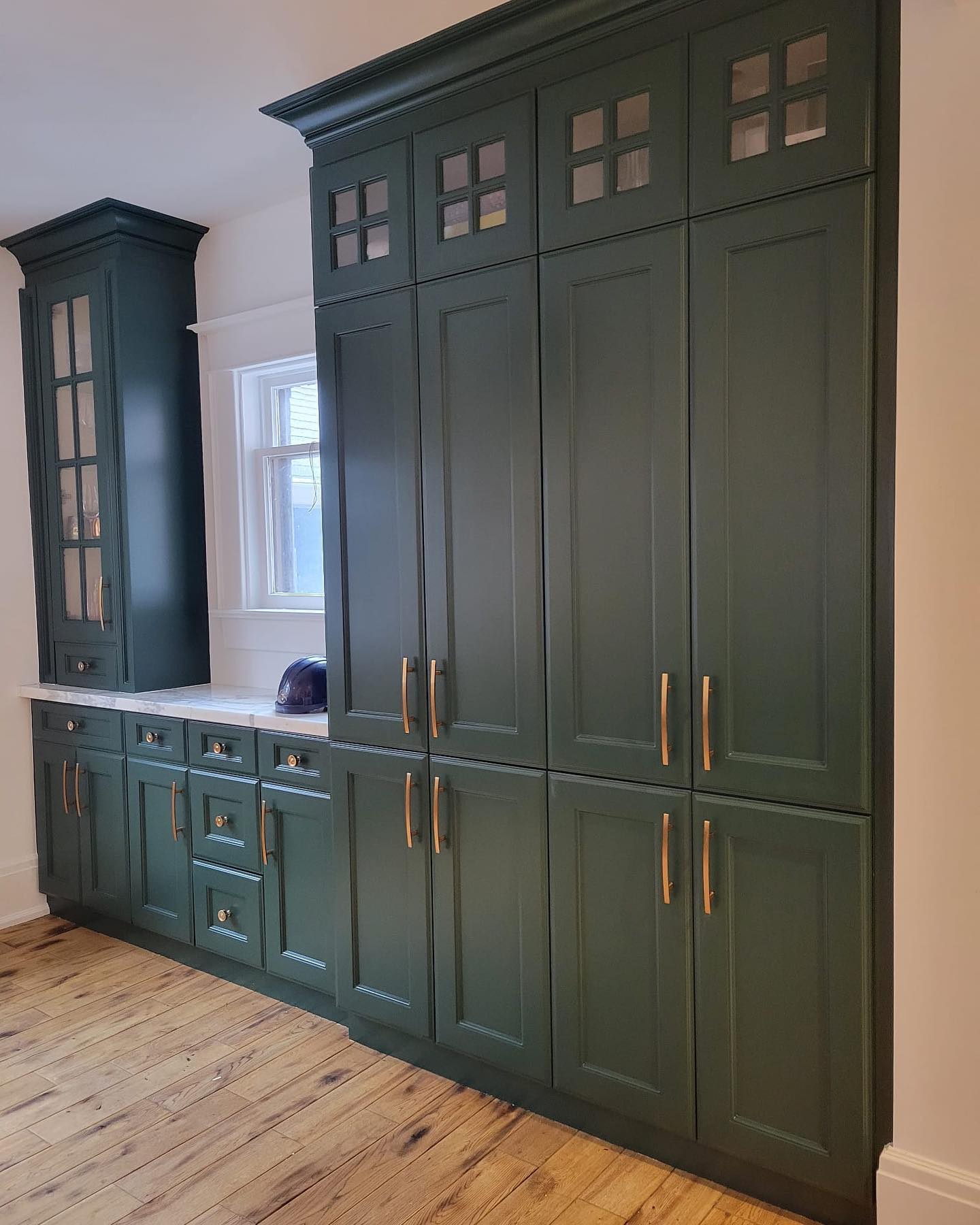 A kitchen with tall, dark green cabinets featuring gold handles, a wooden floor, and a white countertop beneath a window—perfect inspiration for your next home renovation or to share with your favorite contractor.
