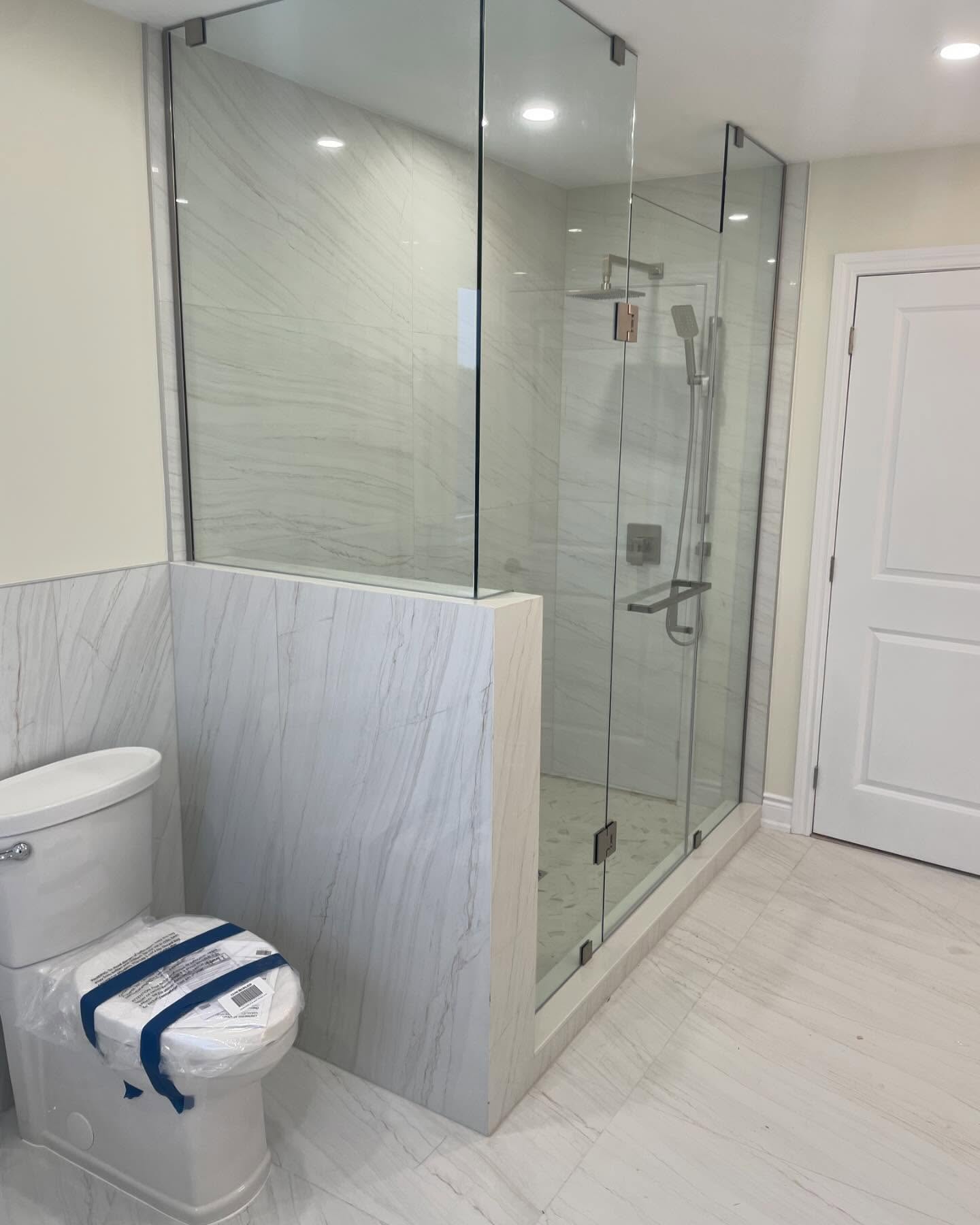 Modern bathroom with white marble tile, designed by Revival Construction, features a glass-enclosed shower with chrome fixtures and built-in bench. A toilet with the lid taped closed sits beside the shower; white walls and a closed door complete this home renovation.