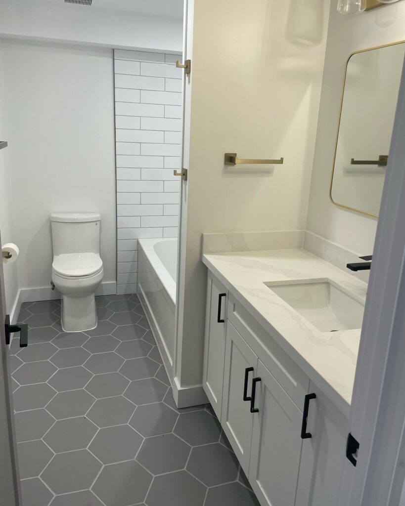 Modern bathroom with gray hexagonal floor tiles, white vanity with a rectangular sink, brass fixtures, a toilet, and a bathtub with white subway tile surround. Perfect for any home renovation by a skilled builder or contractor. Clean, bright look throughout.