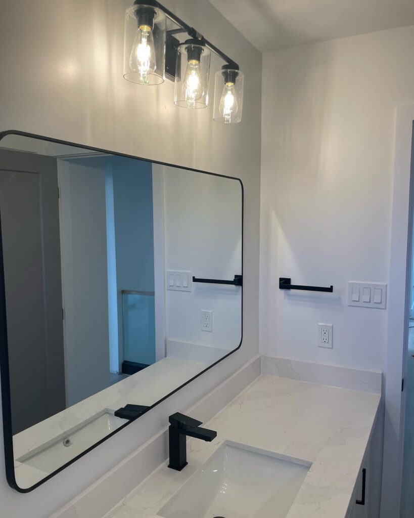 A modern bathroom by Revival Construction features a rectangular mirror, black-framed light fixture with three exposed bulbs, matte black faucet, towel bar, and toilet paper holder, all set against crisp white walls and a sleek white countertop.