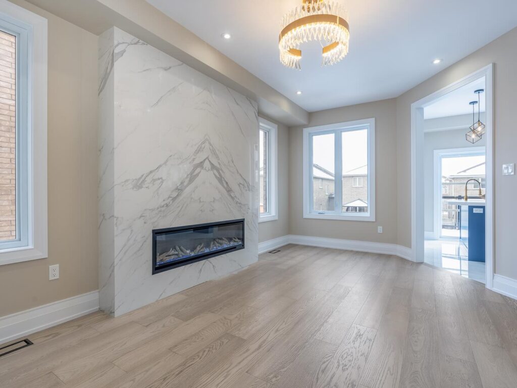 Spacious, modern living room with light wood floors, large windows, a marble fireplace, and a gold chandelier. Sunlight brightens the room, expertly crafted by a skilled builder with an open doorway leading to the kitchen area.