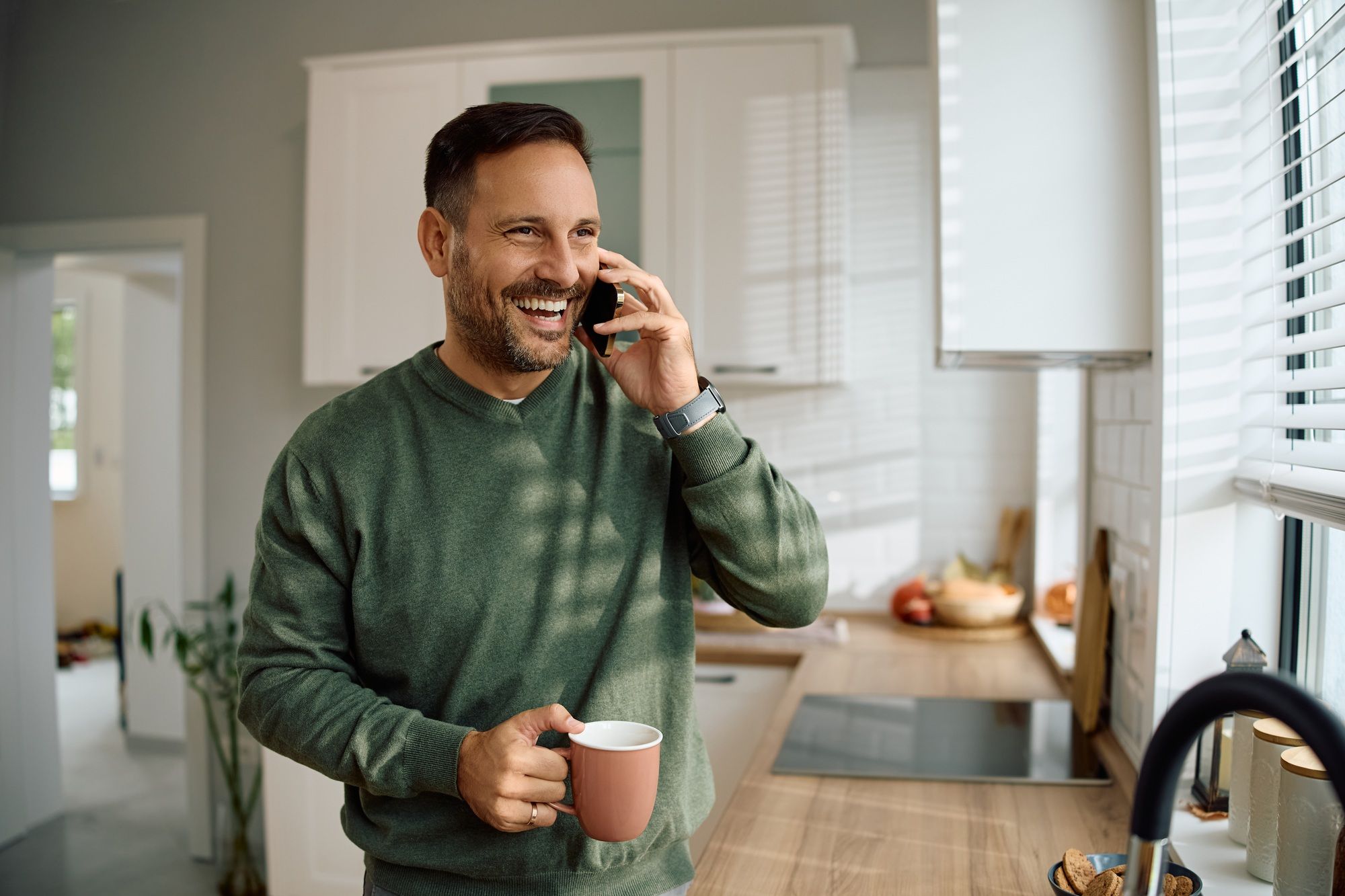 A smiling man in a green sweater stands in a bright kitchen, holding a pink mug and talking on his smartphone, perhaps discussing his renovation plans with Revival Construction while looking out the window.