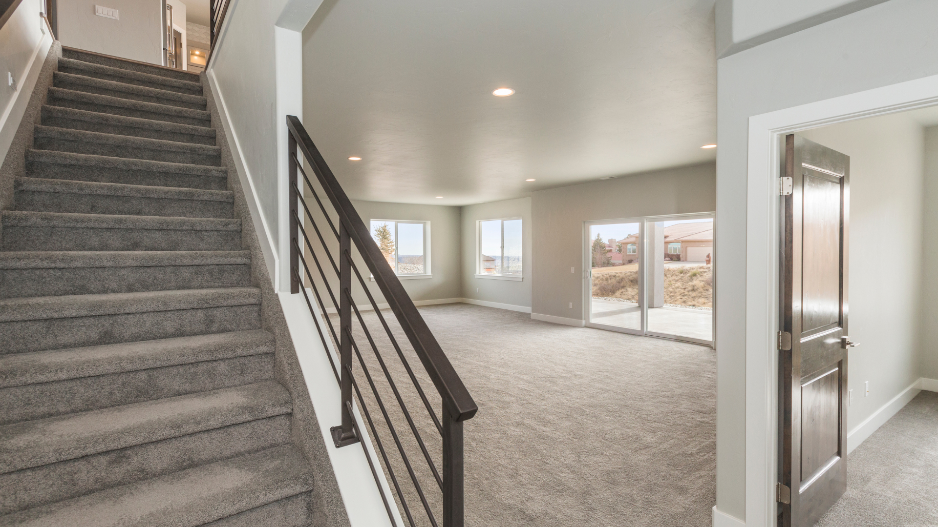 Modern, empty living room with large windows and sliding glass doors, next to a carpeted staircase with a black railing; neutral walls and carpet throughout, with natural light flooding the space.