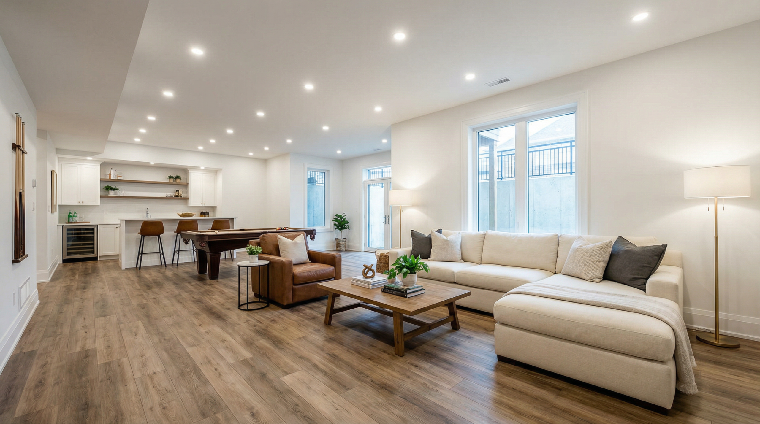 Modern open-concept living space with wood flooring, white sectional sofa, coffee table, armchair, floor lamp, pool table, bar stools, and a kitchen area with white cabinets and sleek lighting.