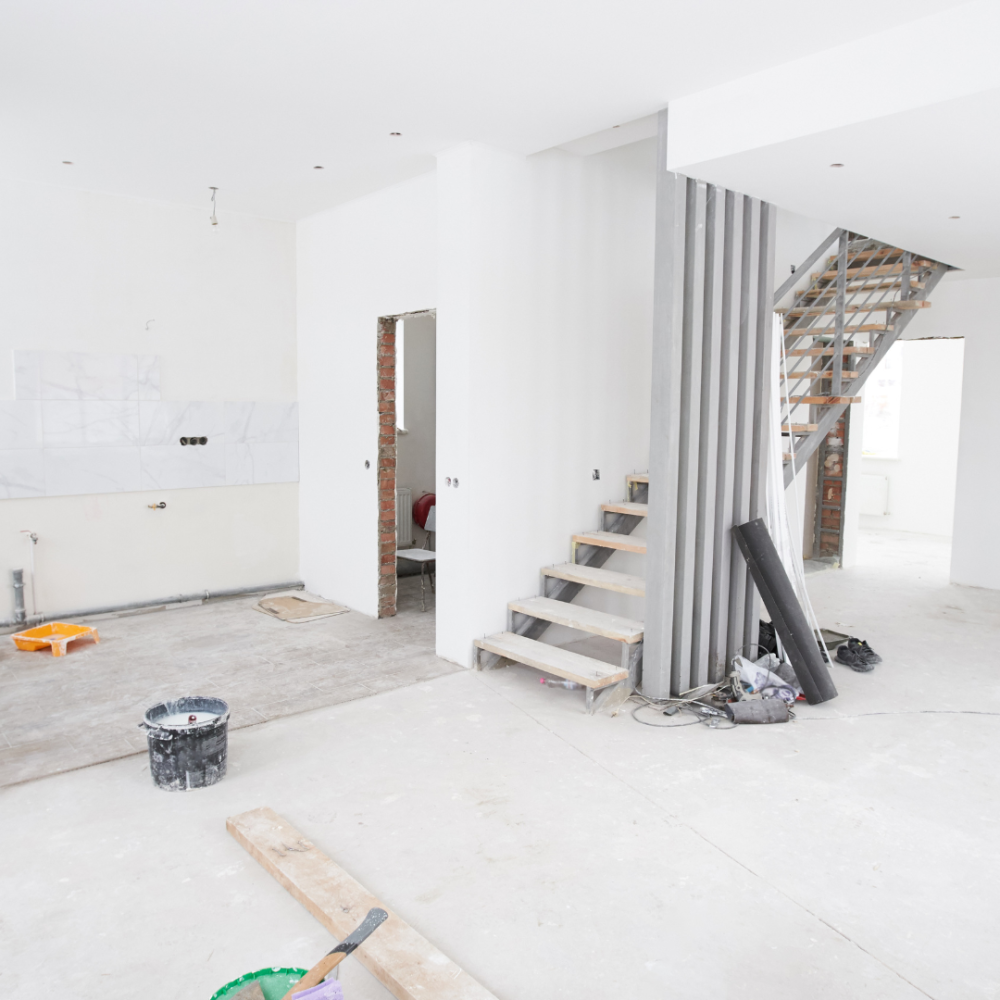 A bright, unfinished interior space with white walls, scattered construction materials, a broom, buckets, a partially completed staircase, and exposed wiring, indicating ongoing renovation or remodeling work.