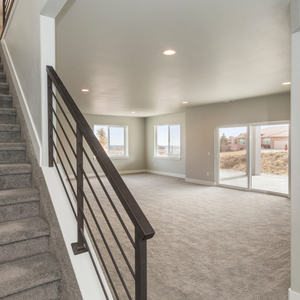 Modern, empty living room with large windows and sliding glass doors, next to a carpeted staircase with a black railing; neutral walls and carpet throughout, with natural light flooding the space.